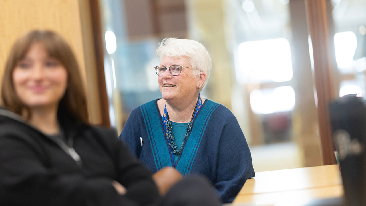 carrie kaas, an older woman with short white hair and glasses, smiles and looks at something off camera. there is a young female student sitting out of focus in front of her