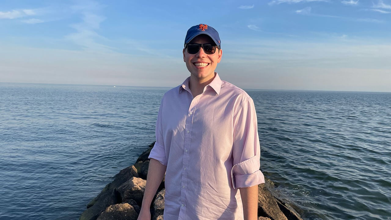 nathanael scaniffe smiles on the beach wearing a baseball cap and sunglasses in front of the water