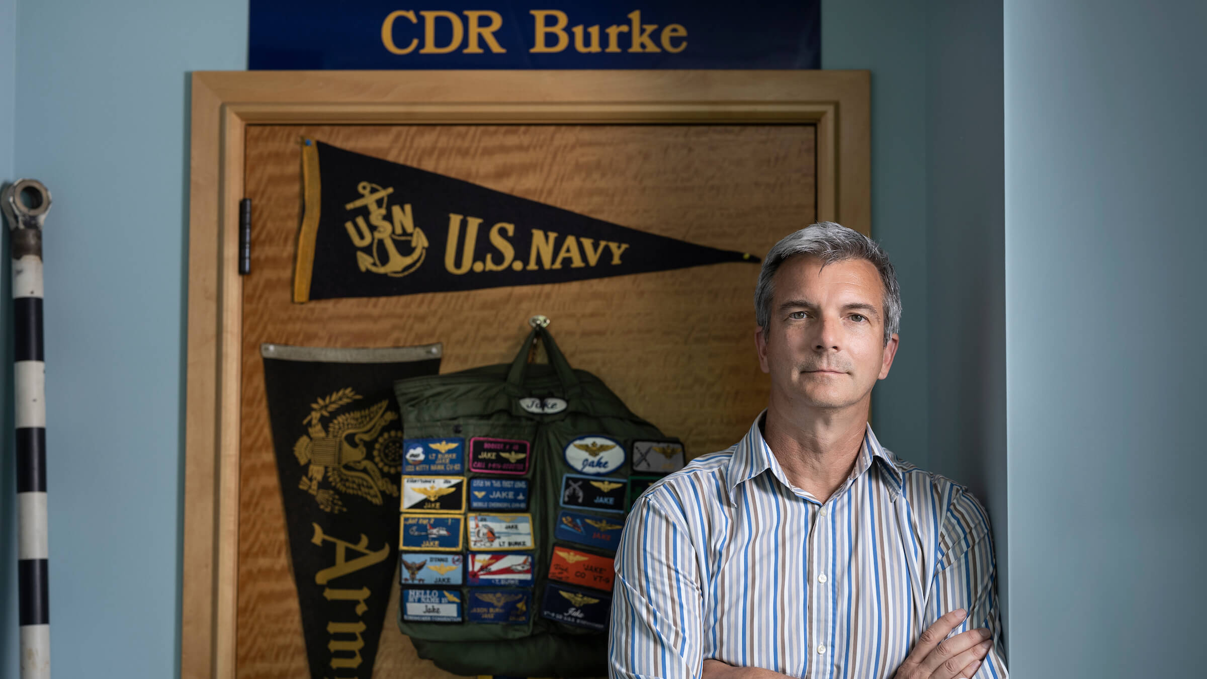 Jason Burke in his home standing in front of US Navy flag and backpack with patches