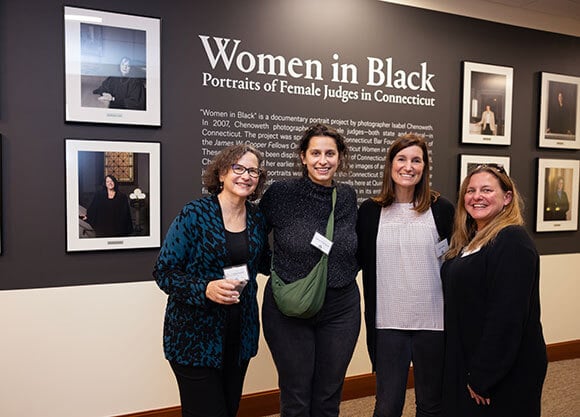 A group of women stand in front of a School of Law exhibit of women judges in Connecticut