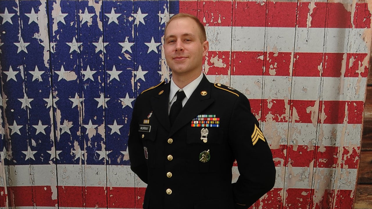 Benjamin Murray in full army uniform standing in front of an american flag.