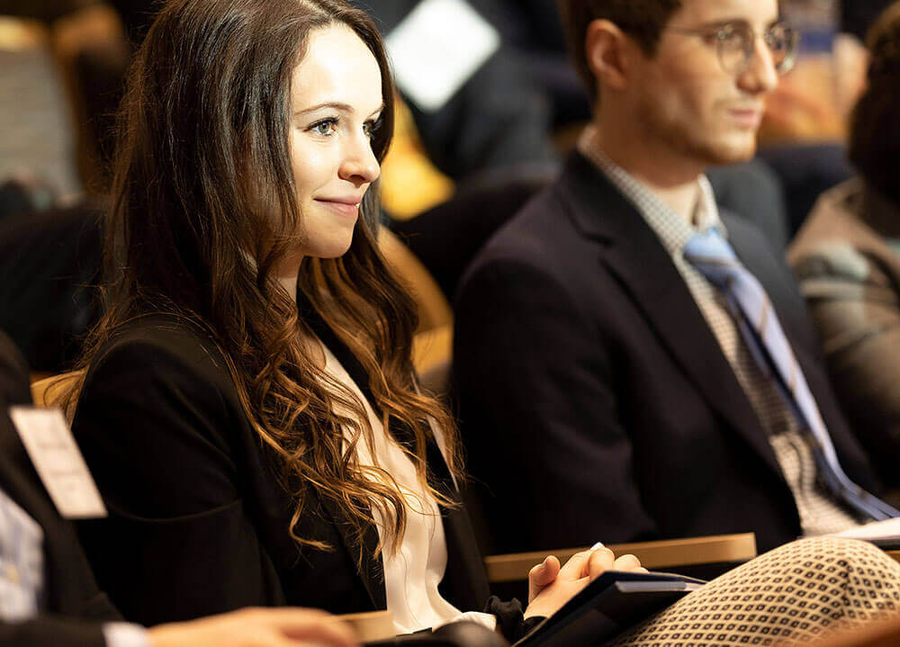 A law student smiles and listens intently to a lecture while sitting in a packed audience in the ceremonial courtroom